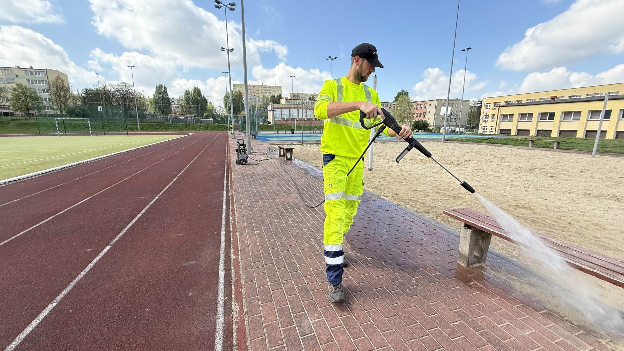 Pracownik myjący ławki myjką ciśnieniową przy bieżni szkolnej — serwis obiektów sportowych Kalisz