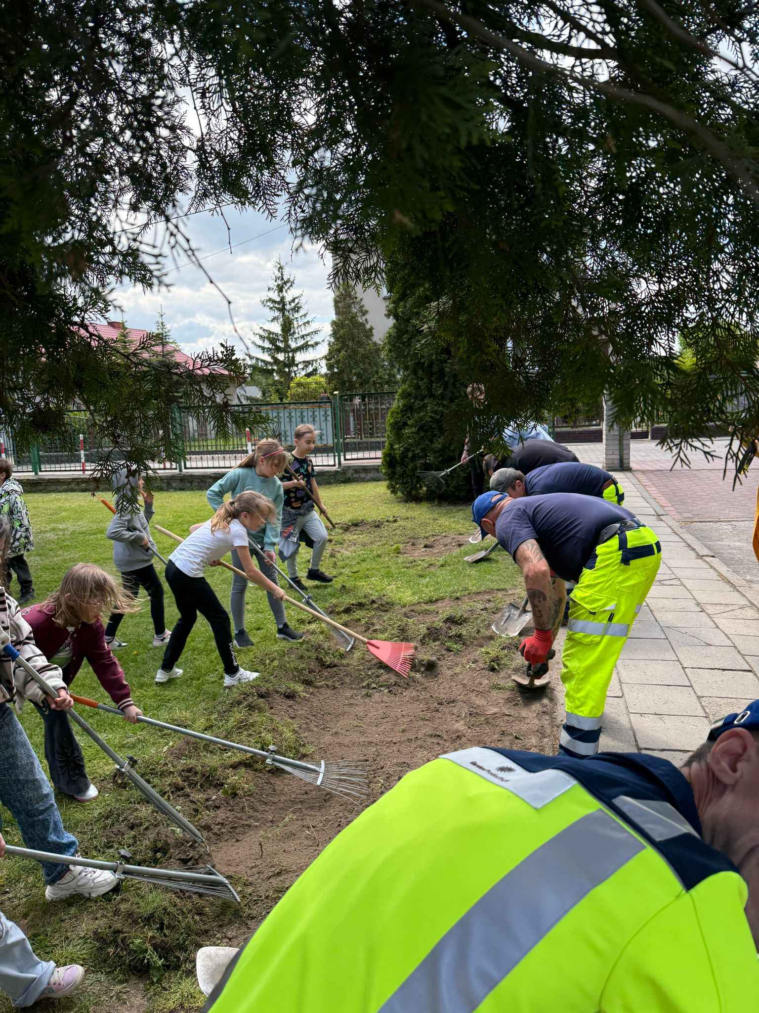 Dzieci grabią rabatę razem z pracownikami GardenProducts — wspólna akcja sadzenia przy szkole