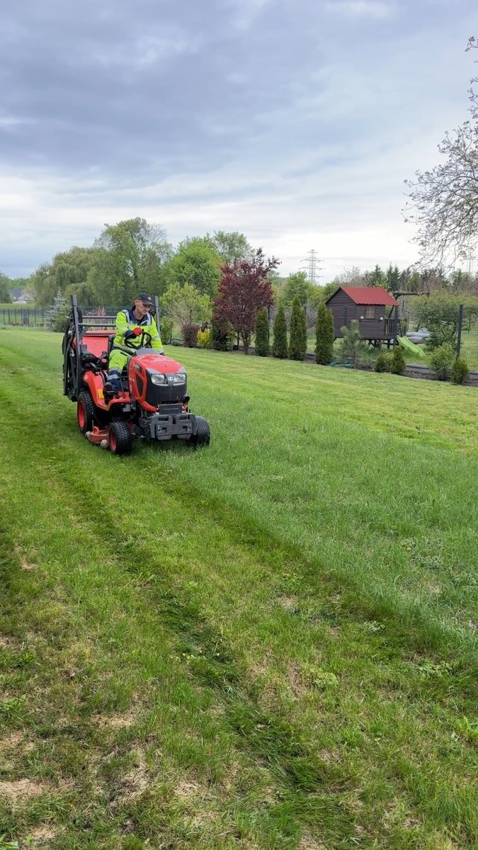 Koszenie dużego trawnika traktorem z koszem zbierającym przy ogrodzie z placem zabaw