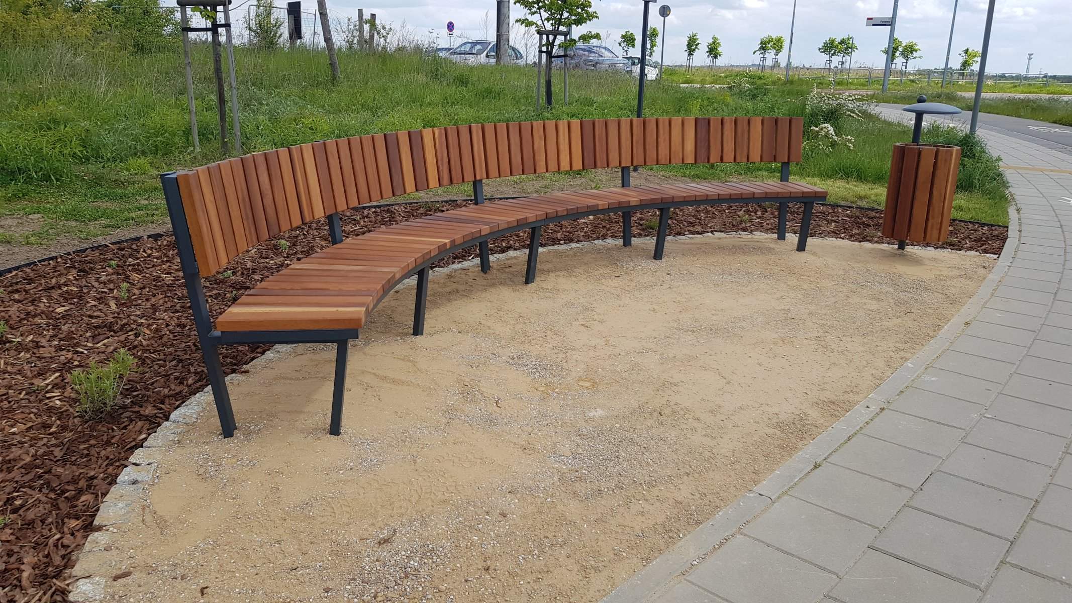Łukowa ławka parkowa z drewna egzotycznego przy ścieżce rowerowej — mała architektura GardenProducts