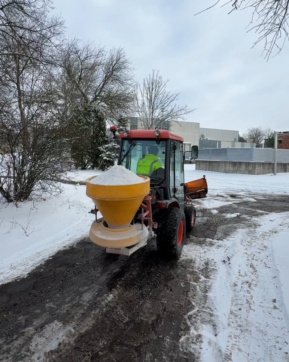 Ciągnik z posypywarką rozprowadza materiał przeciwpoślizgowy — usuwanie gołoledzi solanką i piaskiem